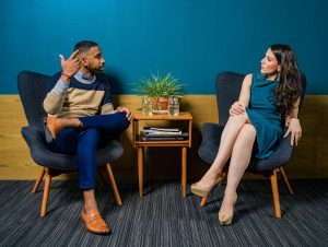 Woman wearing teal dress sitting on chair having conversation with man